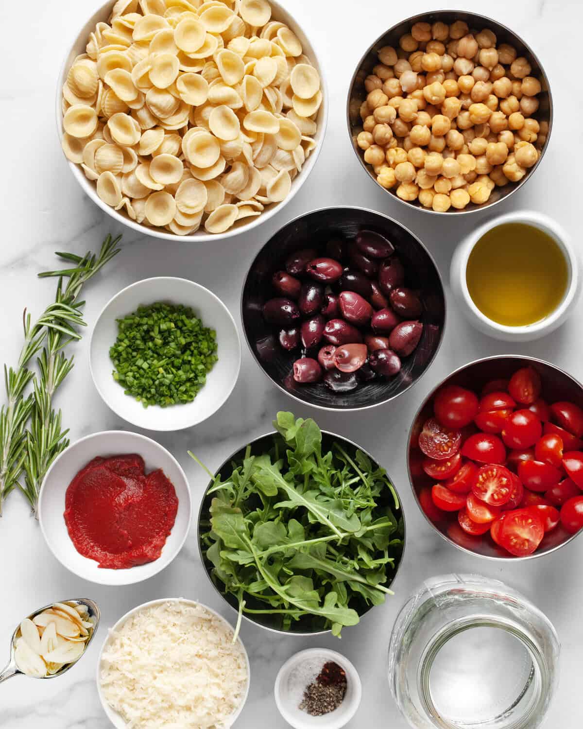 Ingredients including orecchiette, chickpeas, olives, tomatoes, arugula, parmesan, tomato pasta, garlic and water.