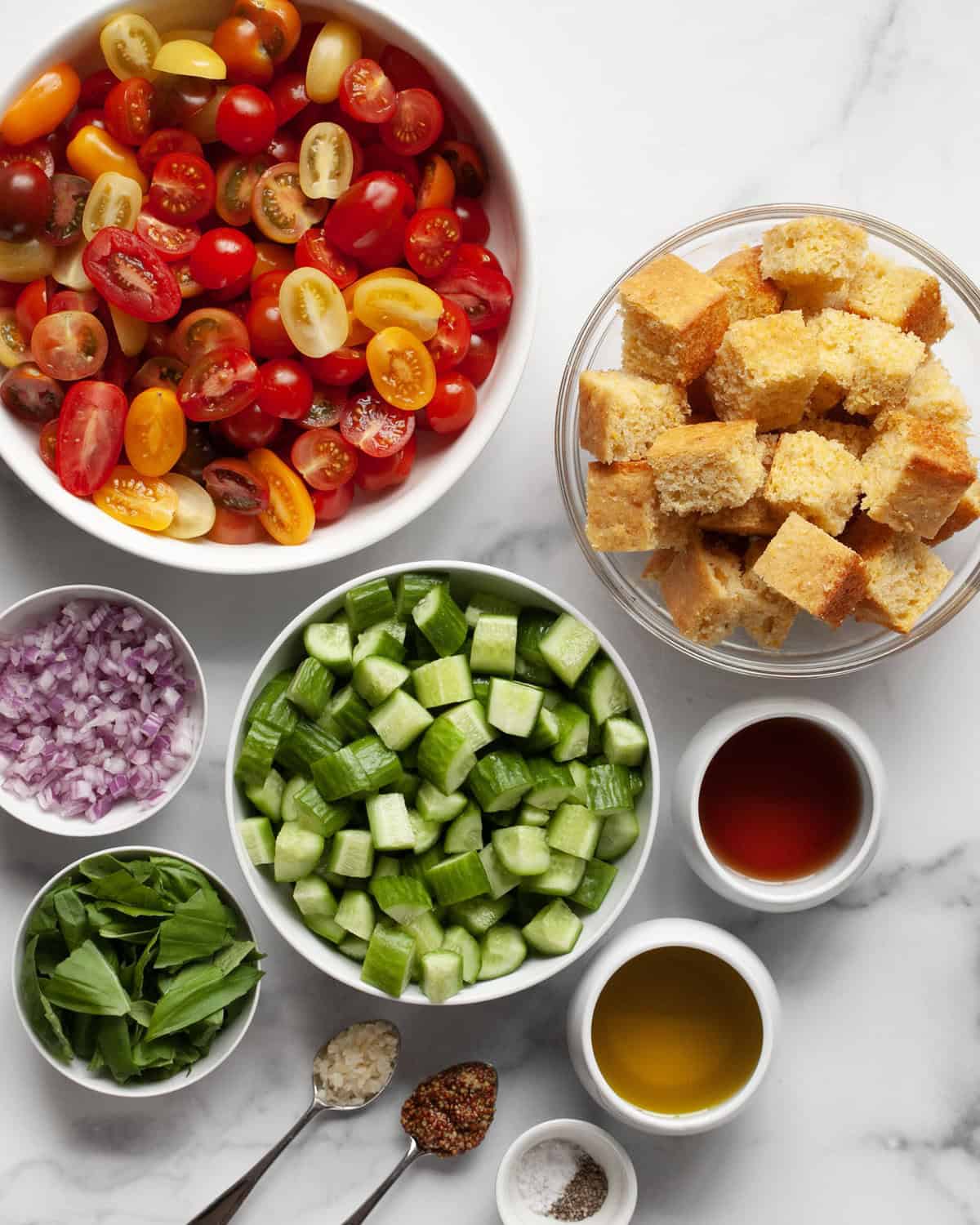Ingredients for salad including cornbread cubes, tomatoes, cucumbers,bers, red onions, basil, vinegar, oil, mustard, garlic, salt and pepper.