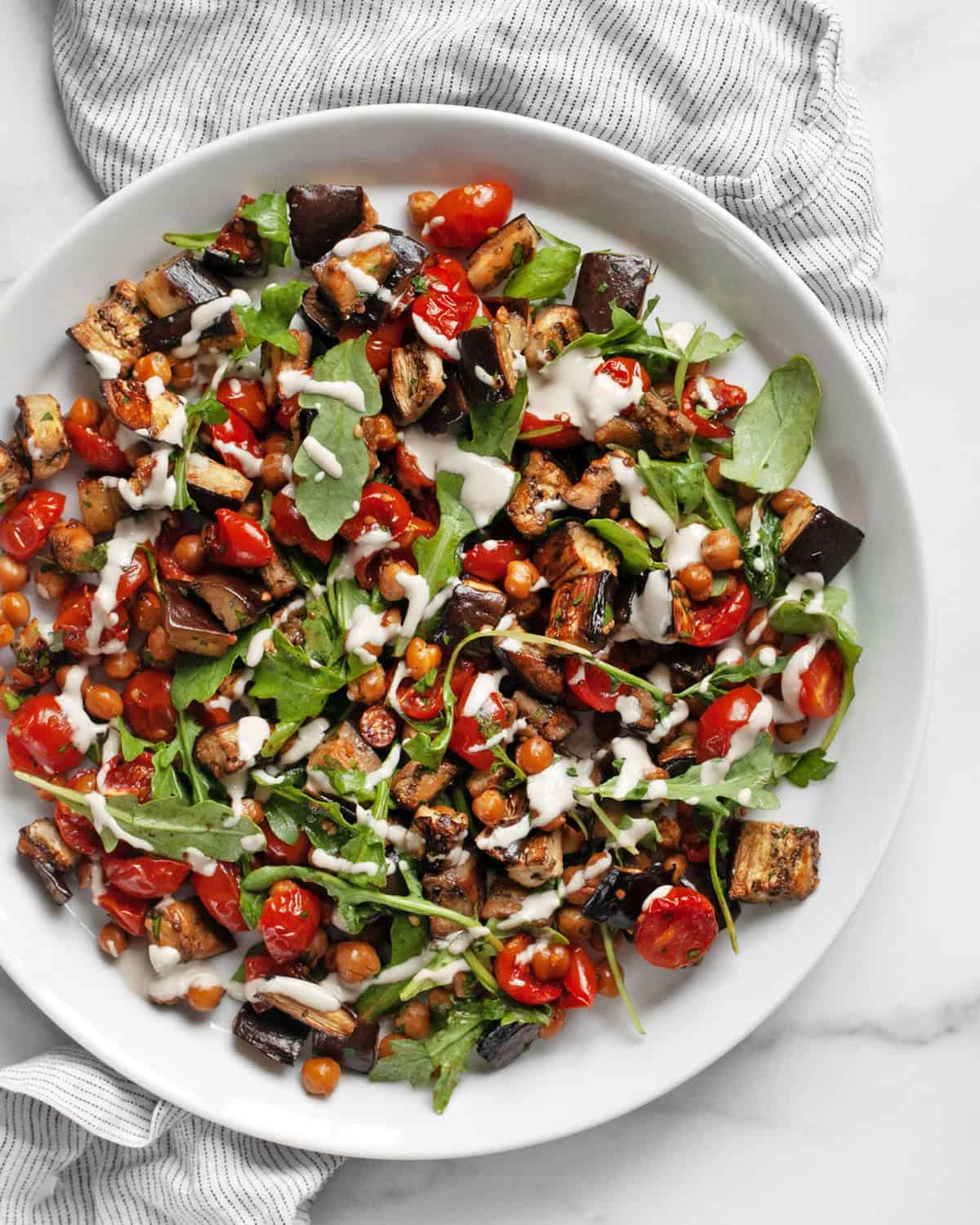 Roasted eggplant, tomatoes and chickpeas  drizzled with lemon tahini sauce on a plate.