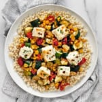 Baked feta with corn, tomatoes and zucchini with pearl couscous on a plate.