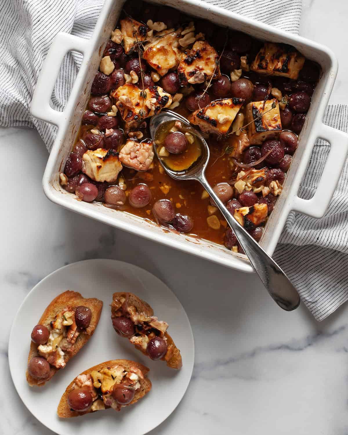 Baked feta and grapes in a dish with a serving spoon and a plate with three pieces of bread topped with grapes and cheese.