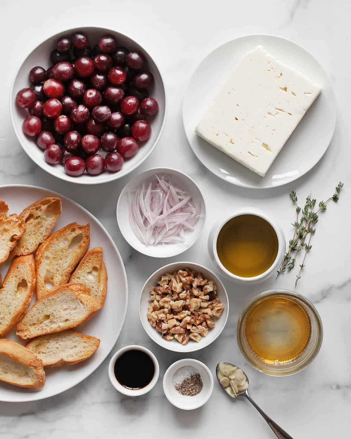 Ingredients including feta, grapes, olive oil, balsamic vinegar, shallots, sliced garlic, thyme, salt, pepper and toasted bread.