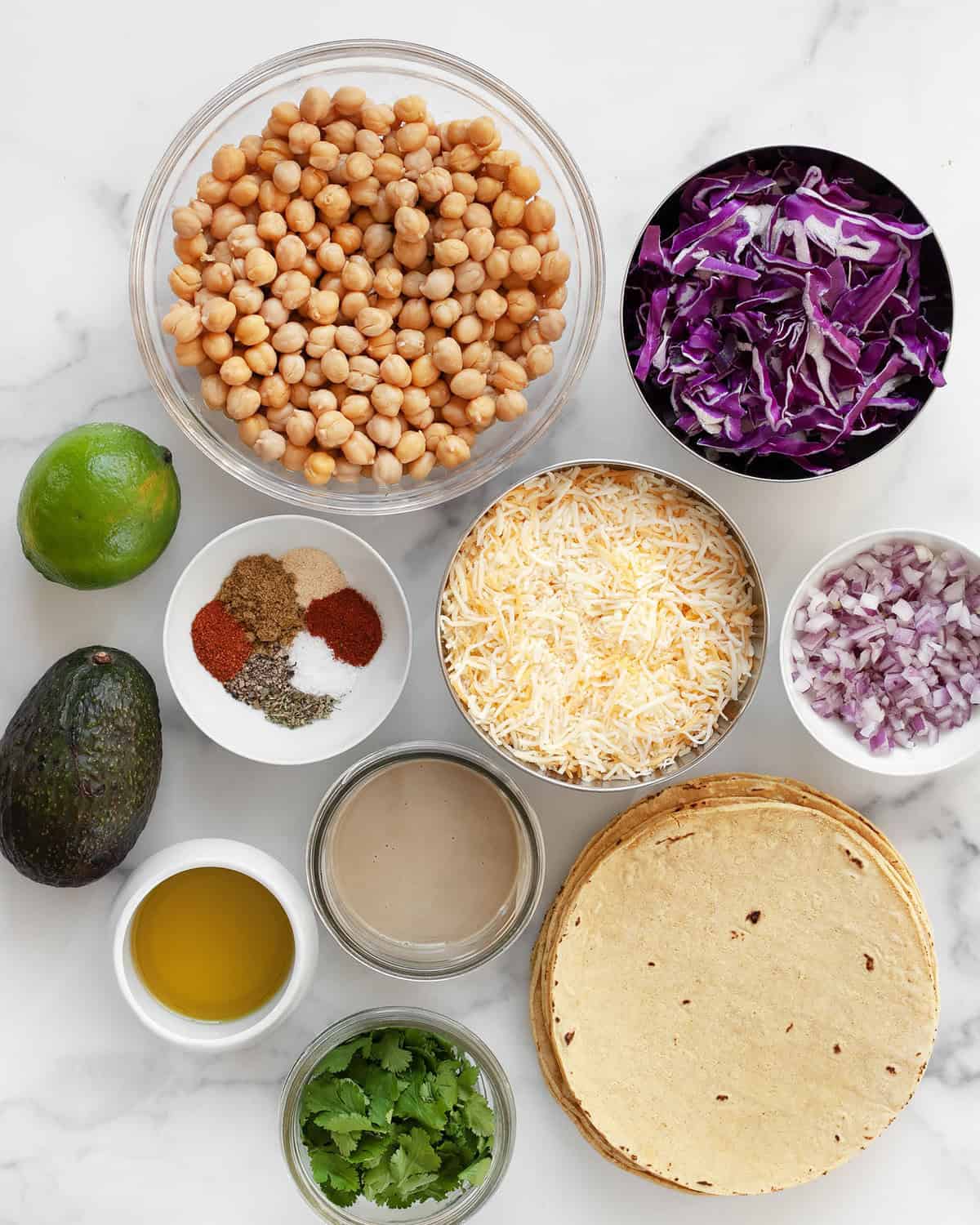 Ingredients including chickpeas, corn tortillas, cabbage, cheese, red onions, taco seasonings, lime, avocado and olive oil.