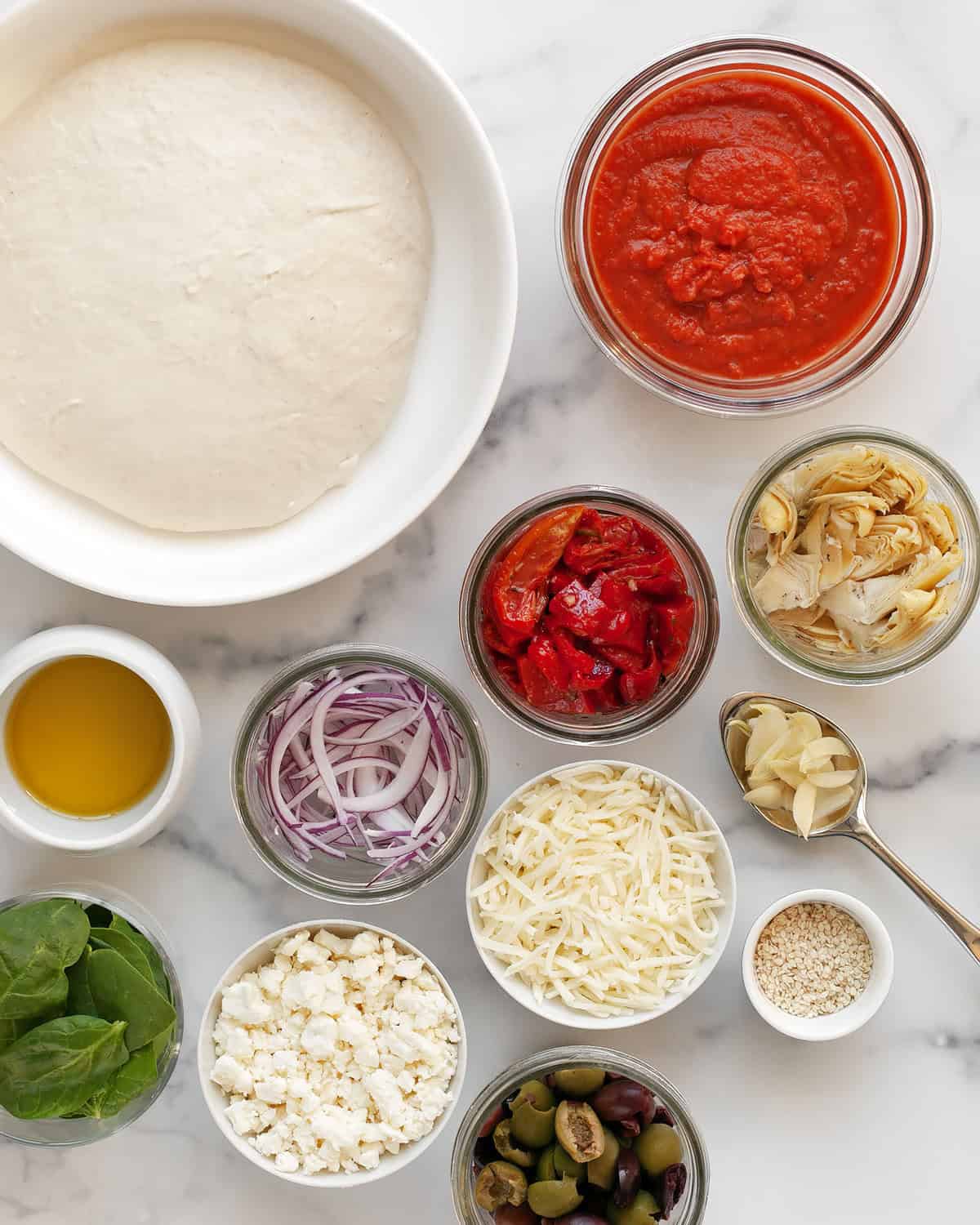 Ingredients including dough, mozzarella, feta, tomato sauce, olives, sun dried tomatoes, artichokes, red peppers, onions, garlic and olive oil.