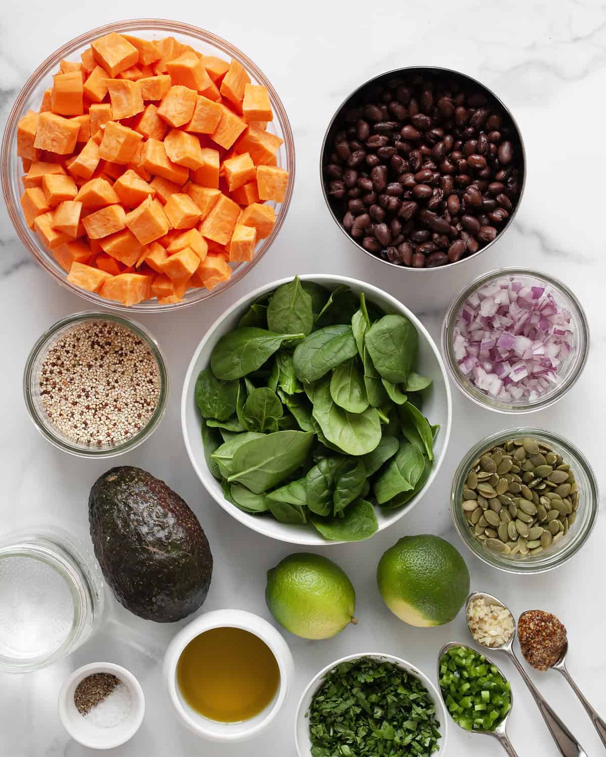 Ingredients including sweet potatoes, black beans, avocado, red onions, quinoa, spinach, pumpkin seeds, limes, garlic, oil and cilantro.