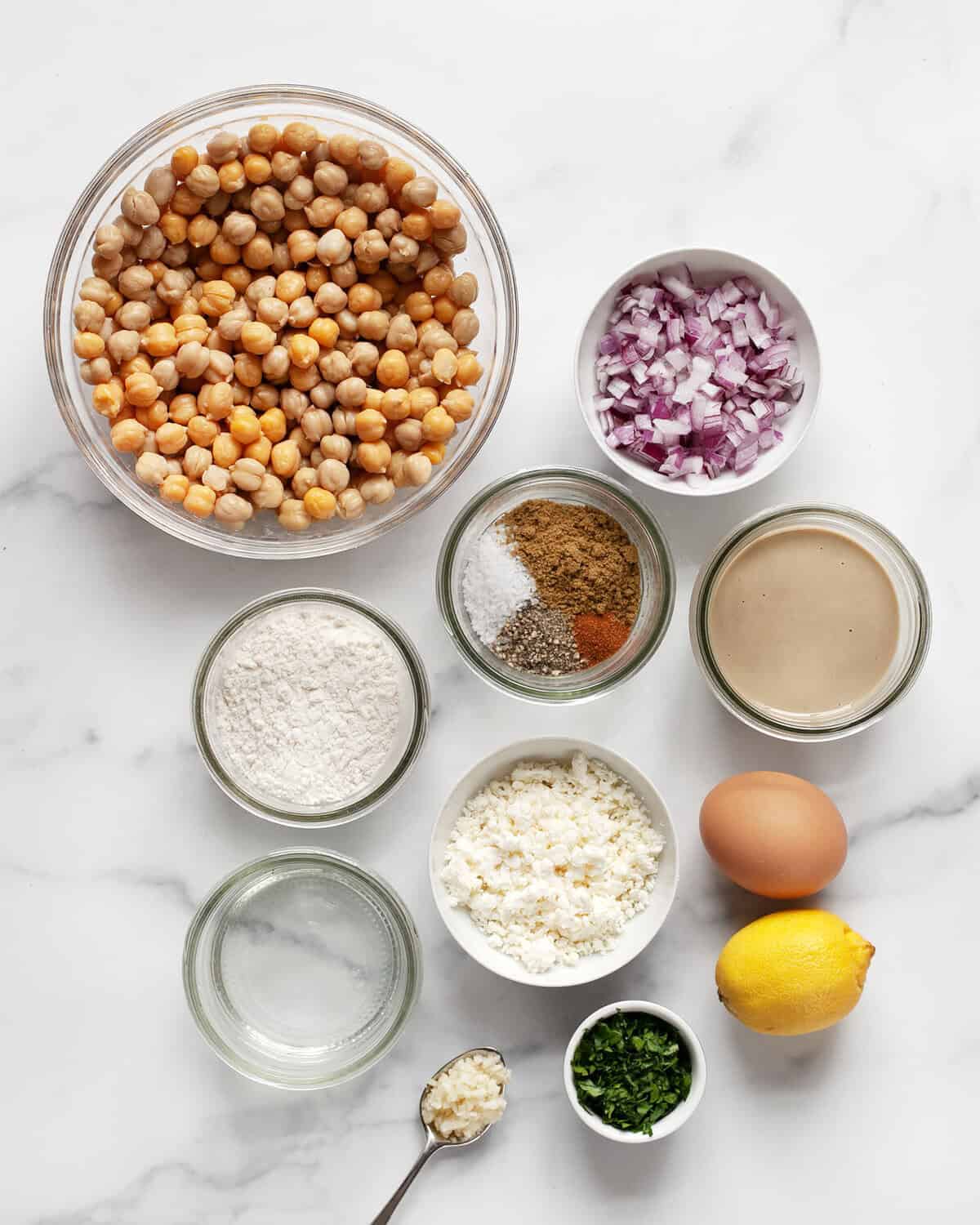 Ingredients including chickpeas, red onions, dried spices, lemon, egg, flour, feta, parsley, garlic, tahini and water.